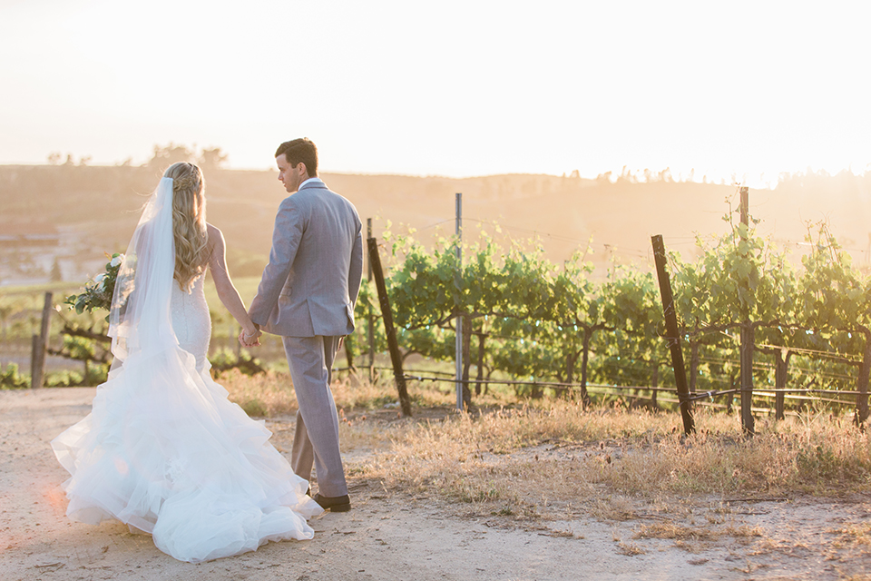 Temecula outdoor wedding at falkner winery bride mermaid style gown with lace bodice and sweetheart neckline with ruffled skirt and long veil with groom heather grey suit with white dress shirt and long white tie with matching pocket square and white floral boutonniere walking and holding hands