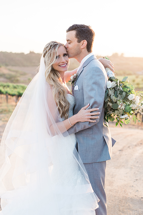 Temecula outdoor wedding at falkner winery bride mermaid style gown with lace bodice and sweetheart neckline with ruffled skirt and long veil with groom heather grey suit with white dress shirt and long white tie with matching pocket square and white floral boutonniere hugging and kissing bride holding white and green floral bridal bouquet