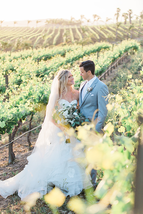 Temecula outdoor wedding at falkner winery bride mermaid style gown with lace bodice and sweetheart neckline with ruffled skirt and long veil with groom heather grey suit with white dress shirt and long white tie with matching pocket square and white floral boutonniere hugging in vineyard bride holding white and green floral bridal bouquet