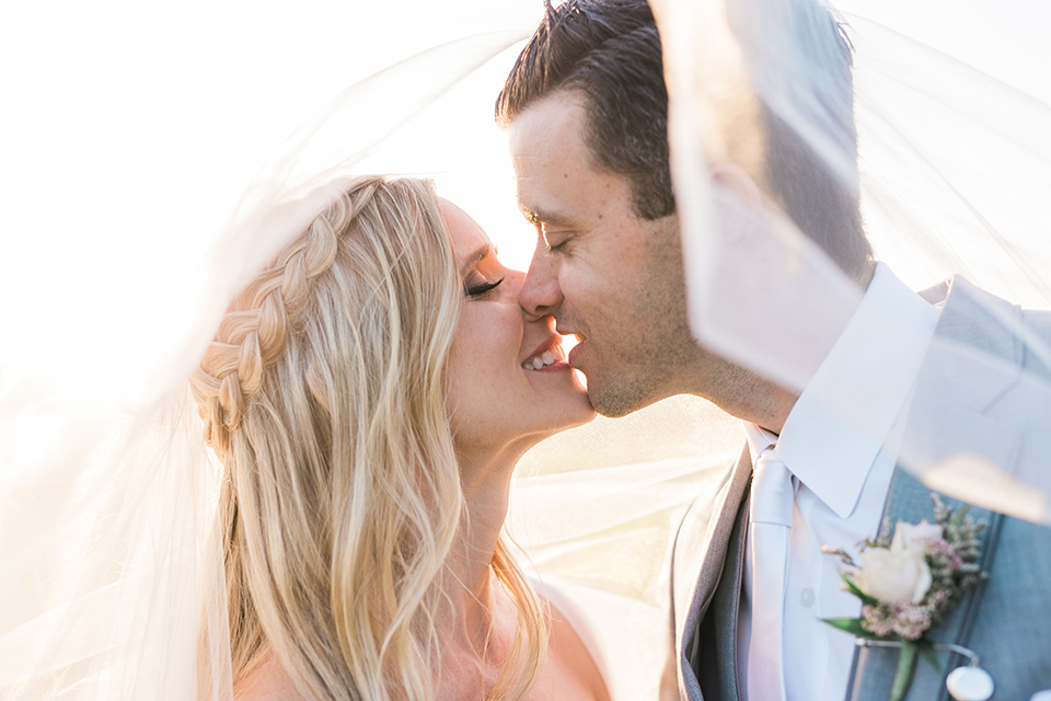 Temecula outdoor wedding at falkner winery bride mermaid style gown with lace bodice and sweetheart neckline with ruffled skirt and long veil with groom heather grey suit with white dress shirt and long white tie with matching pocket square and white floral boutonniere kissing under veil