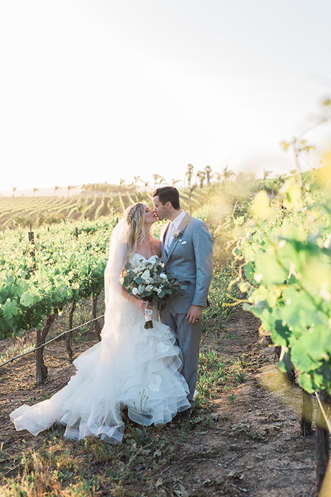 Temecula outdoor wedding at falkner winery bride mermaid style gown with lace bodice and sweetheart neckline with ruffled skirt and long veil with groom heather grey suit with white dress shirt and long white tie with matching pocket square and white floral boutonniere kissing in vineyard bride holding white and green floral bridal bouquet