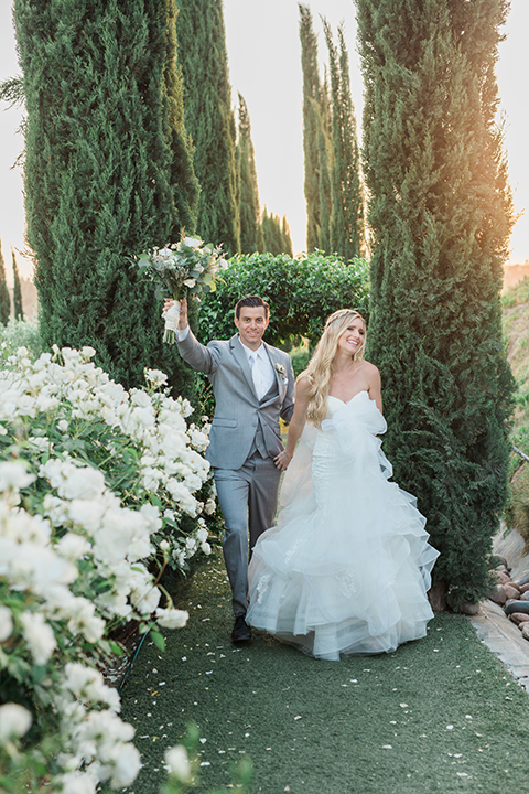 Temecula outdoor wedding at falkner winery bride mermaid style gown with lace bodice and sweetheart neckline with ruffled skirt and long veil with groom heather grey suit with white dress shirt and long white tie with matching pocket square and white floral boutonniere walking and holding hands groom holding brides white and green floral bridal bouquet