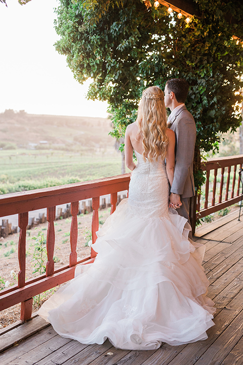 Temecula outdoor wedding at falkner winery bride mermaid style gown with lace bodice and sweetheart neckline with ruffled skirt and long veil with groom heather grey suit with white dress shirt and long white tie with matching pocket square and white floral boutonniere holding hands on balcony