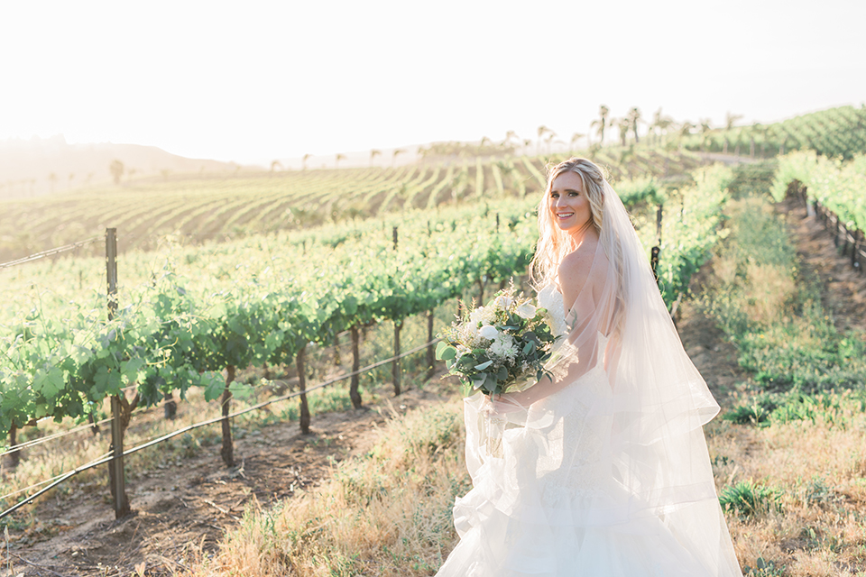 Temecula outdoor wedding at falkner winery bride mermaid style gown with lace bodice and sweetheart neckline with ruffled skirt and long veil holding white and green floral bridal bouquet in vineyard