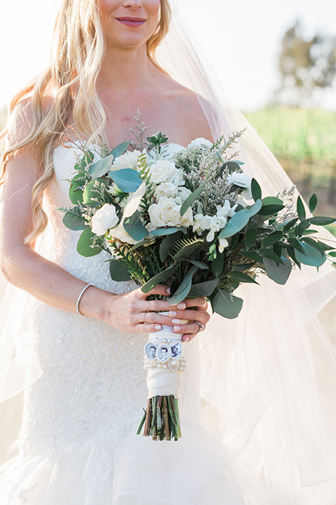 Temecula outdoor wedding at falkner winery bride mermaid style gown with lace bodice and sweetheart neckline with ruffled skirt and long veil holding white and green floral bridal bouquet in vineyard close up