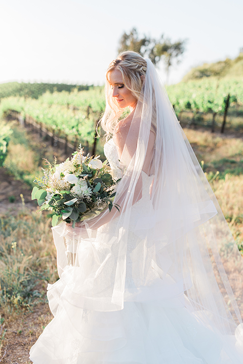 Temecula outdoor wedding at falkner winery bride mermaid style gown with lace bodice and sweetheart neckline with ruffled skirt and long veil holding white and green floral bridal bouquet in vineyard