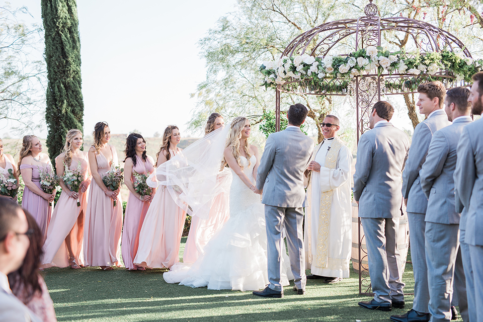 Temecula outdoor wedding at falkner winery bride mermaid style gown with lace bodice and sweetheart neckline with ruffled skirt and long veil with groom heather grey suit with white dress shirt and long white tie with matching pocket square and white floral boutonniere holding hands during ceremony