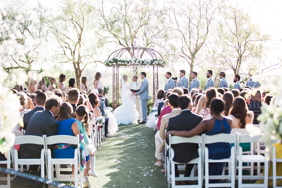 Temecula outdoor wedding at falkner winery bride mermaid style gown with lace bodice and sweetheart neckline with ruffled skirt and long veil with groom heather grey suit with white dress shirt and long white tie with matching pocket square and white floral boutonniere holding hands during ceremony