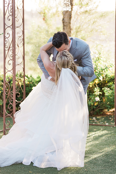 Temecula outdoor wedding at falkner winery bride mermaid style gown with lace bodice and sweetheart neckline with ruffled skirt and long veil with groom heather grey suit with white dress shirt and long white tie with matching pocket square and white floral boutonniere kissing during ceremony