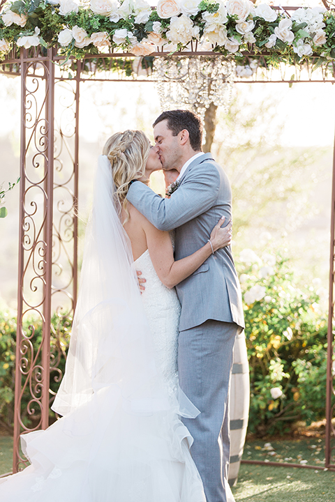 Temecula outdoor wedding at falkner winery bride mermaid style gown with lace bodice and sweetheart neckline with ruffled skirt and long veil with groom heather grey suit with white dress shirt and long white tie with matching pocket square and white floral boutonniere kissing during ceremony