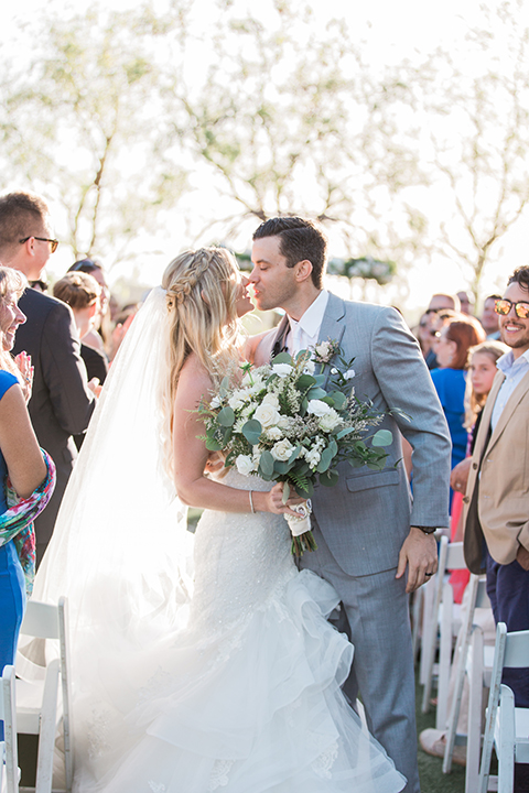 Temecula outdoor wedding at falkner winery bride mermaid style gown with lace bodice and sweetheart neckline with ruffled skirt and long veil with groom heather grey suit with white dress shirt and long white tie with matching pocket square and white floral boutonniere kissing after ceremony bride holding white and green floral bridal bouquet