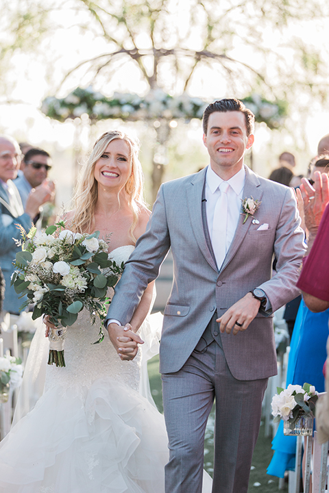 Temecula outdoor wedding at falkner winery bride mermaid style gown with lace bodice and sweetheart neckline with ruffled skirt and long veil with groom heather grey suit with white dress shirt and long white tie with matching pocket square and white floral boutonniere holding hands and walking down aisle after ceremony bride holding white and green floral bridal bouquet