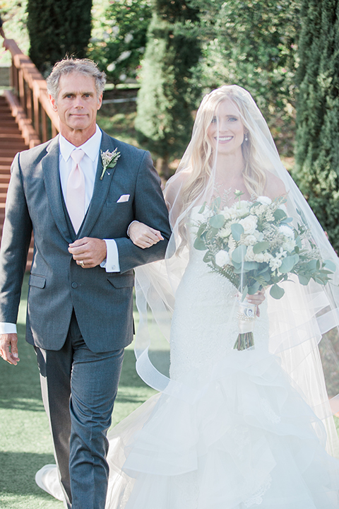 Temecula outdoor wedding at falkner winery bride mermaid style gown with lace bodice and sweetheart neckline with ruffled skirt and long veil holding white and green floral bridal bouquet in vineyard walking down the aisle with dad veil over head