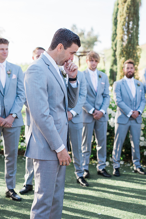 Temecula outdoor wedding at falkner winery groom heather grey suit with matching vest and white dress shirt with long white tie and matching pocket square with white floral boutonniere crying watching bride walk down the aisle