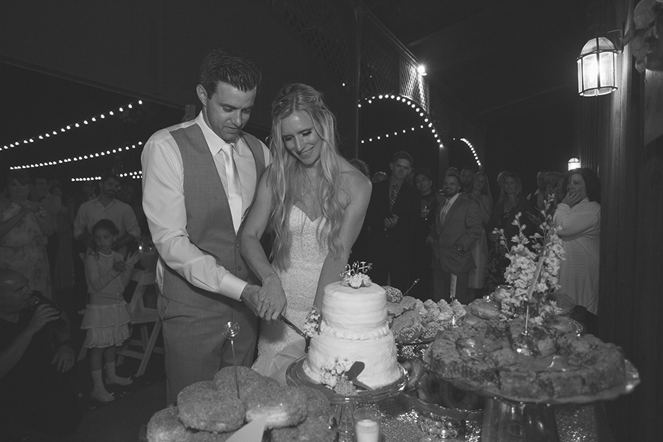 Temecula outdoor wedding at falkner winery bride mermaid style gown with lace bodice and sweetheart neckline with ruffled skirt and long veil with groom heather grey suit with white dress shirt and long white tie with matching pocket square and white floral boutonniere cutting cake at reception