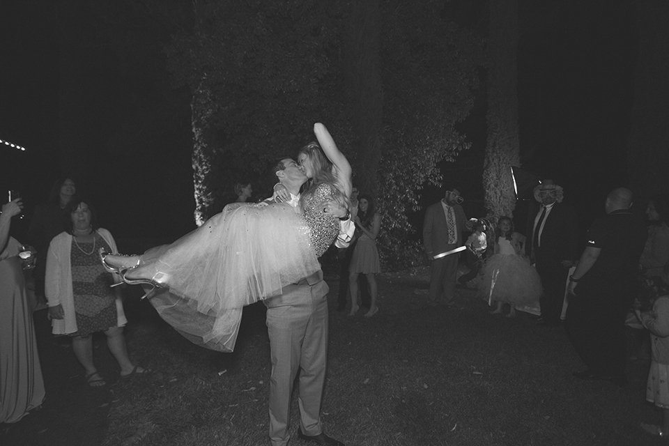 Temecula outdoor wedding at falkner winery bride mermaid style gown with lace bodice and sweetheart neckline with ruffled skirt and long veil with groom heather grey suit with white dress shirt and long white tie with matching pocket square and white floral boutonniere kissing at reception black and white photo