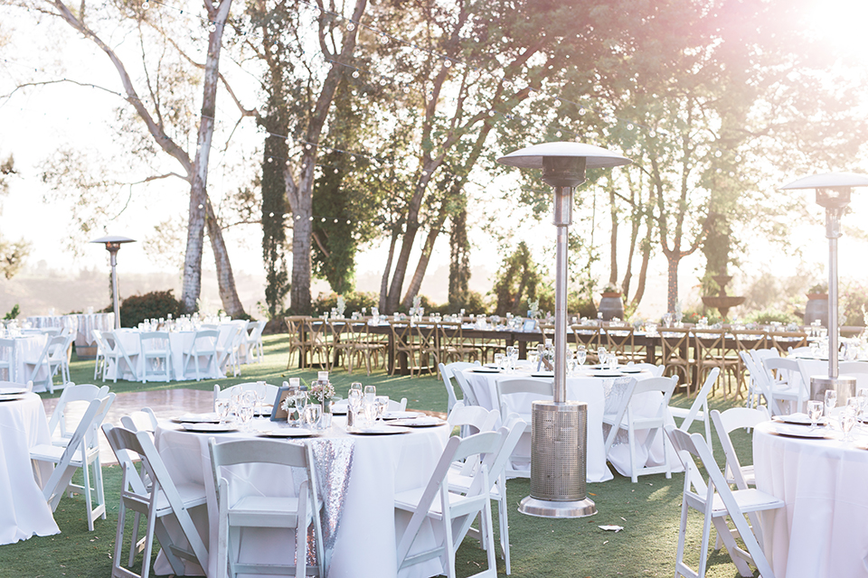 Temecula outdoor wedding at falkner winery reception set up with tables white table linen with white chairs and white place settings with pink and white name place and white flower decor with long light brown wood table with matching wood chairs and flower decor