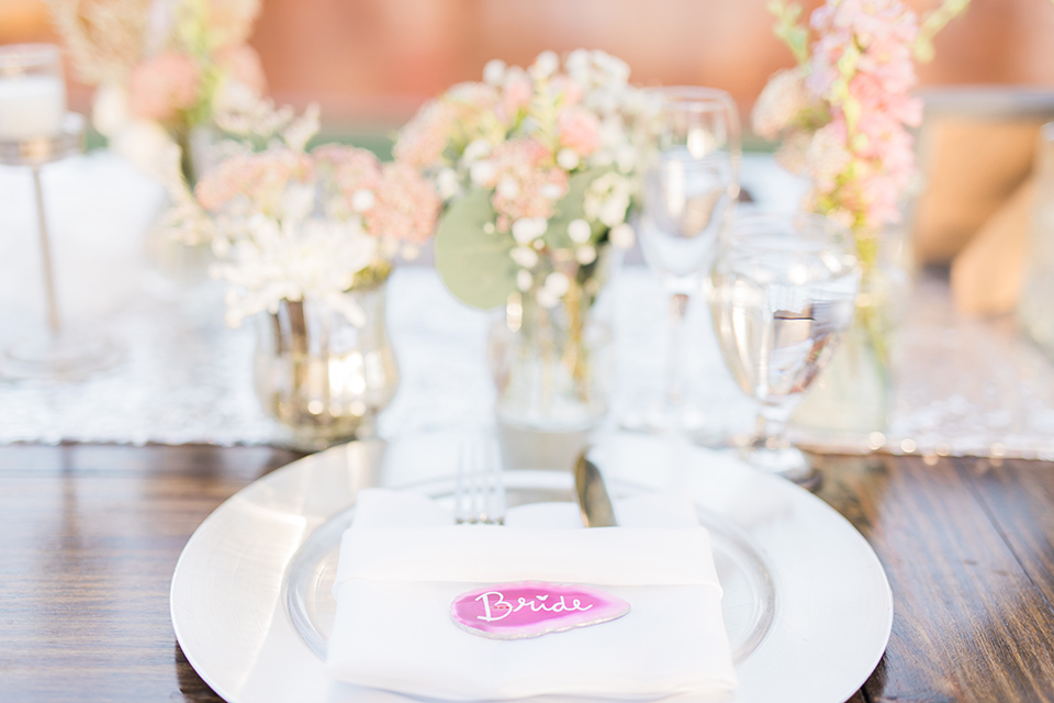 Temecula outdoor wedding at falkner winery reception set up with tables white table linen with white chairs and white place settings with pink and white name place and white flower decor with long light brown wood table with matching wood chairs and flower decor