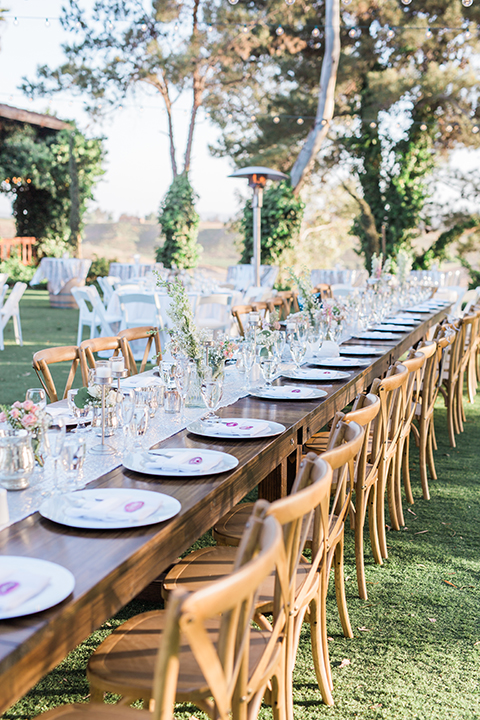 Temecula outdoor wedding at falkner winery reception set up with tables white table linen with white chairs and white place settings with pink and white name place and white flower decor with long light brown wood table with matching wood chairs and flower decor