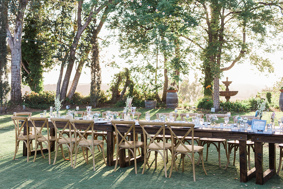 Temecula outdoor wedding at falkner winery reception set up with tables white table linen with white chairs and white place settings with pink and white name place and white flower decor with long light brown wood table with matching wood chairs and flower decor