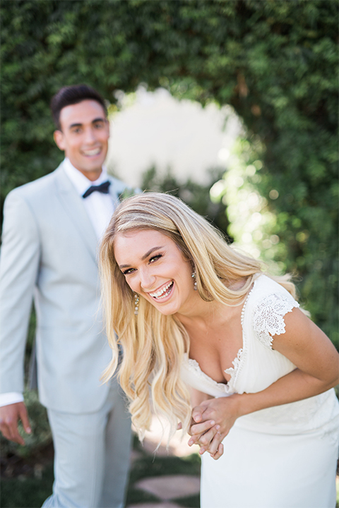 Orange county outdoor wedding at balboa bay resort bride form fitting lace gown with plunging neckline and lace straps with groom light grey peak lapel suit with matching vest and white dress shirt with charcoal grey bow tie and green floral boutonniere laughing