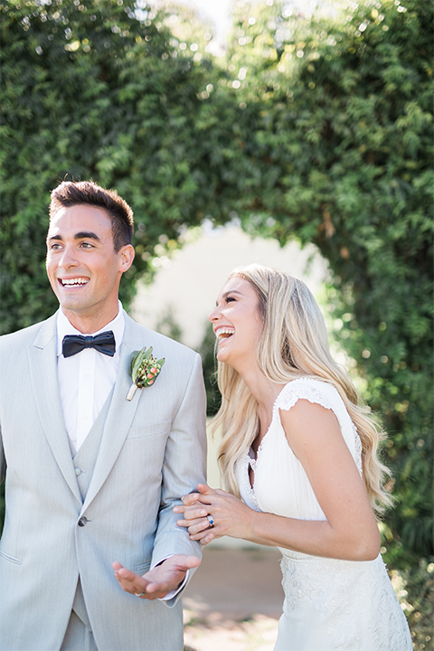 Balboa Bay Resort Wedding Bride form fitting lace gown with plunging neckline and lace straps and Groom light grey peak lapel suit with matching vest and white dress shirt with charcoal bow tie and green floral boutonniere Laughing