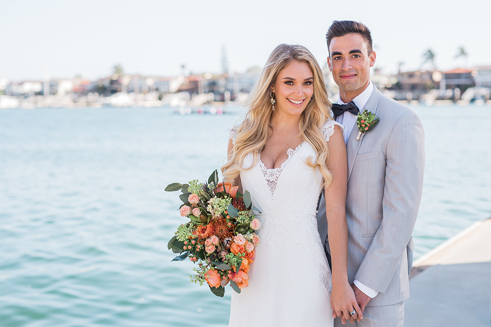 Balboa Bay Resort Wedding Bride and Groom Standing by Water Holding Flowers Grey Suit Navy Bow Tie Brown Shoes