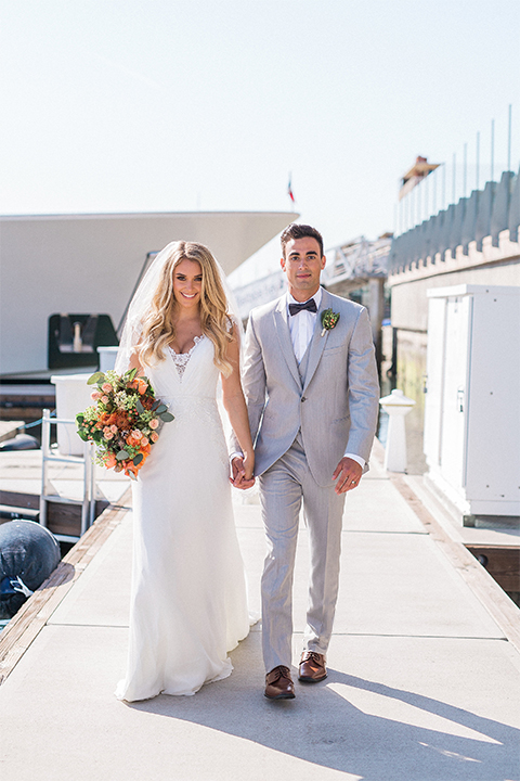 Balboa Bay Resort Wedding Bride and Groom Walking on Dock Holding Flowers Grey Suit Navy Bow Tie Brown Shoes