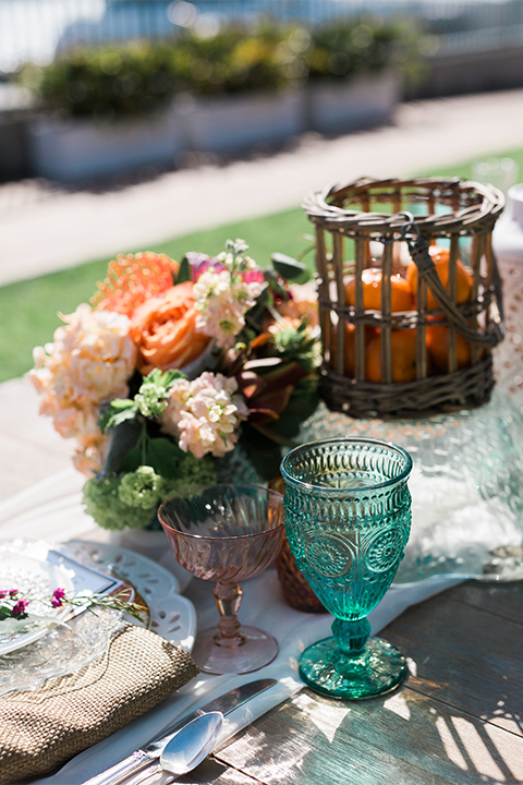 Orange county outdoor wedding at balboa bay resort table set up with light brown wood table with white lace table runner and white place settings with blue glasses and orange flower centerpiece decor 