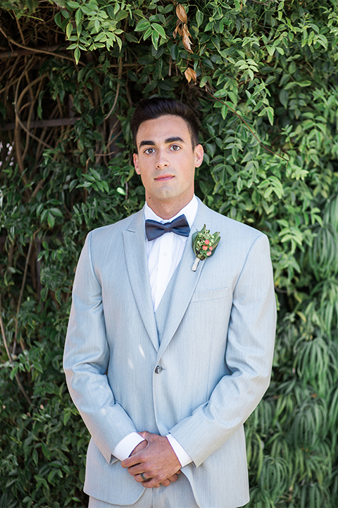 Orange county outdoor wedding at balboa bay resort groom light grey peak lapel suit with matching vest and white dress shirt with charcoal grey bow tie and green floral boutonniere standing with hands crossed
