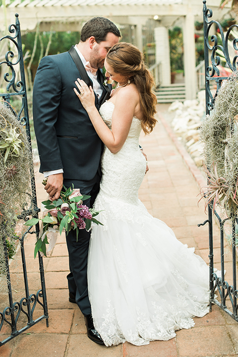 Orange county outdoor wedding at sherman library and gardens bride form fitting strapless lace gown with sweetheart neckline and pearl necklace with groom navy blue shawl lapel tuxedo with matching vest and white dress shirt with blush pink bow tie and white floral boutonniere hugging