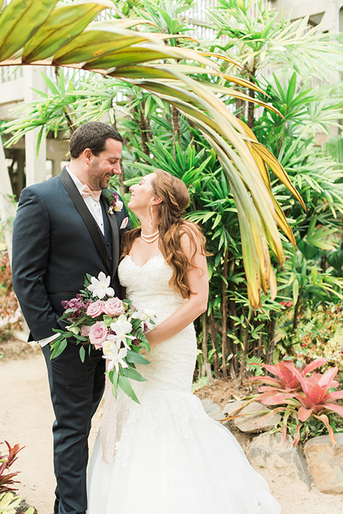 Orange county outdoor wedding at sherman library and gardens bride form fitting strapless lace gown with sweetheart neckline and pearl necklace with groom navy blue shawl lapel tuxedo with matching vest and white dress shirt with blush pink bow tie and white floral boutonniere hugging and bride holding white and pink floral bridal bouquet