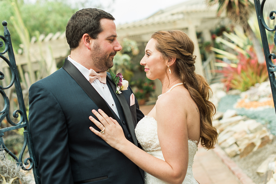 Orange county outdoor wedding at sherman library and gardens bride form fitting strapless lace gown with sweetheart neckline and pearl necklace with groom navy blue shawl lapel tuxedo with matching vest and white dress shirt with blush pink bow tie and white floral boutonniere hugging