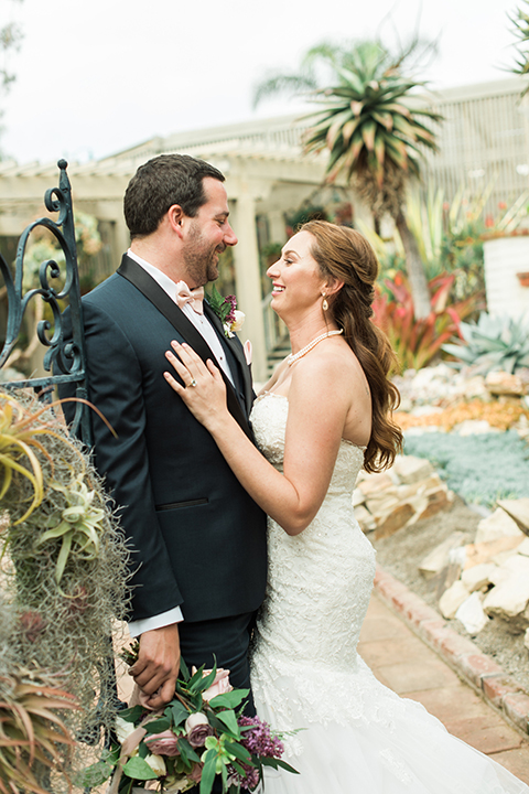 Orange county outdoor wedding at sherman library and gardens bride form fitting strapless lace gown with sweetheart neckline and pearl necklace with groom navy blue shawl lapel tuxedo with matching vest and white dress shirt with blush pink bow tie and white floral boutonniere hugging and groom holding white and pink floral bridal bouquet