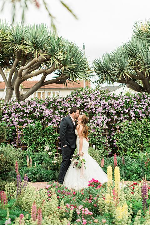Orange county outdoor wedding at sherman library and gardens bride form fitting strapless lace gown with sweetheart neckline and pearl necklace with groom navy blue shawl lapel tuxedo with matching vest and white dress shirt with blush pink bow tie and white floral boutonniere kissing and bride holding white and pink floral bridal bouquet
