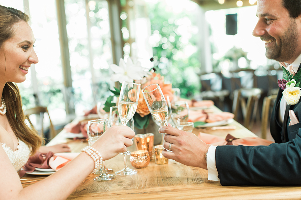 Orange county outdoor wedding at sherman library and gardens bride form fitting strapless lace gown with sweetheart neckline and pearl necklace with groom navy blue shawl lapel tuxedo with matching vest and white dress shirt with blush pink bow tie and white floral boutonniere sitting at table holding drinks