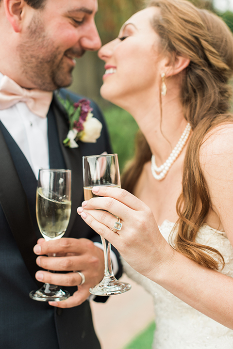 Orange county outdoor wedding at sherman library and gardens bride form fitting strapless lace gown with sweetheart neckline and pearl necklace with groom navy blue shawl lapel tuxedo with matching vest and white dress shirt with blush pink bow tie and white floral boutonniere standing holding drinks