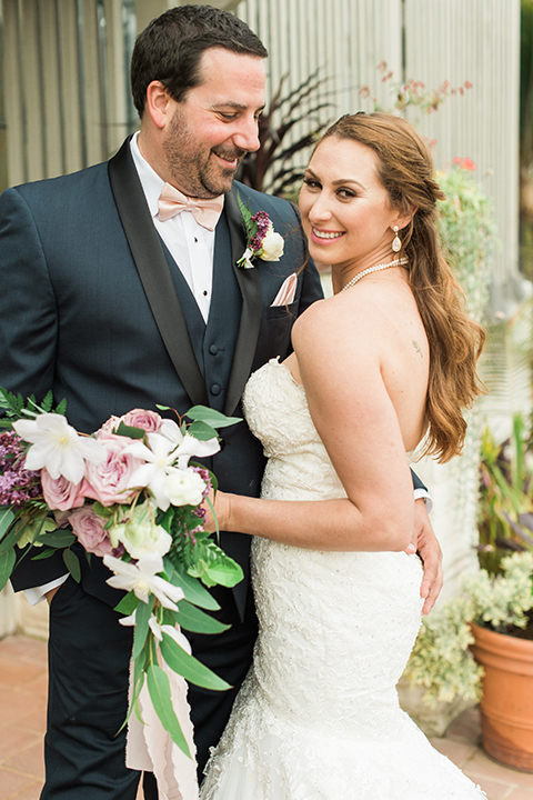 Orange county outdoor wedding at sherman library and gardens bride form fitting strapless lace gown with sweetheart neckline and pearl necklace with groom navy blue shawl lapel tuxedo with matching vest and white dress shirt with blush pink bow tie and white floral boutonniere hugging and groom holding white and pink floral bridal bouquet