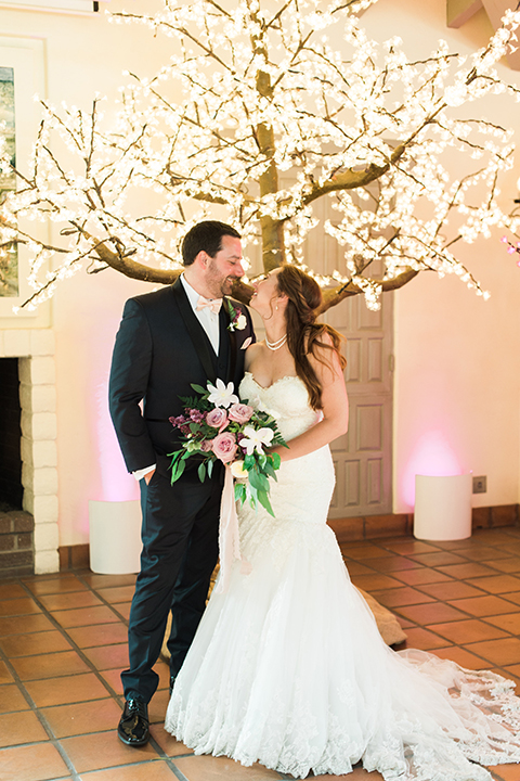 Orange county outdoor wedding at sherman library and gardens bride form fitting strapless lace gown with sweetheart neckline and pearl necklace with groom navy blue shawl lapel tuxedo with matching vest and white dress shirt with blush pink bow tie and white floral boutonniere hugging and bride holding white and pink floral bridal bouquet