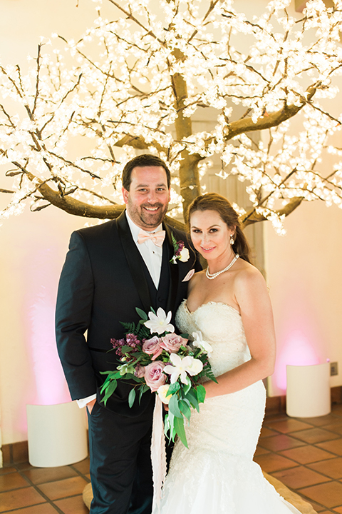 Orange county outdoor wedding at sherman library and gardens bride form fitting strapless lace gown with sweetheart neckline and pearl necklace with groom navy blue shawl lapel tuxedo with matching vest and white dress shirt with blush pink bow tie and white floral boutonniere hugging and bride holding white and pink floral bridal bouquet