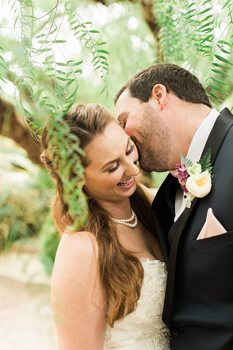 Orange county outdoor wedding at sherman library and gardens bride form fitting strapless lace gown with sweetheart neckline and pearl necklace with groom navy blue shawl lapel tuxedo with matching vest and white dress shirt with blush pink bow tie and white floral boutonniere kissing and bride holding white and pink floral bridal bouquet