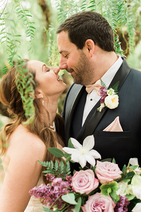 Orange county outdoor wedding at sherman library and gardens bride form fitting strapless lace gown with sweetheart neckline and pearl necklace with groom navy blue shawl lapel tuxedo with matching vest and white dress shirt with blush pink bow tie and white floral boutonniere hugging and bride holding white and pink floral bridal bouquet