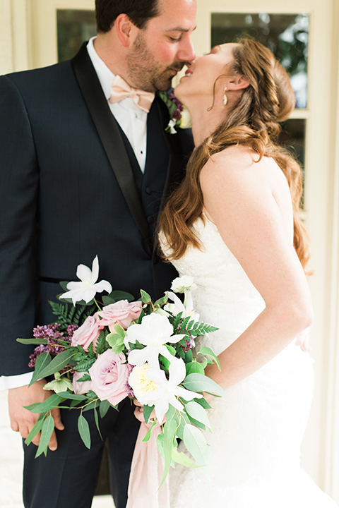 Orange county outdoor wedding at sherman library and gardens bride form fitting strapless lace gown with sweetheart neckline and pearl necklace with groom navy blue shawl lapel tuxedo with matching vest and white dress shirt with blush pink bow tie and white floral boutonniere hugging and bride holding white and pink floral bridal bouquet