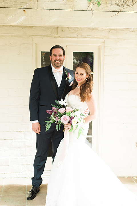 Orange county outdoor wedding at sherman library and gardens bride form fitting strapless lace gown with sweetheart neckline and pearl necklace with groom navy blue shawl lapel tuxedo with matching vest and white dress shirt with blush pink bow tie and white floral boutonniere hugging and bride holding white and pink floral bridal bouquet