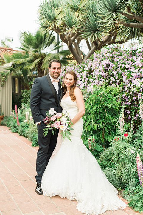 Orange county outdoor wedding at sherman library and gardens bride form fitting strapless lace gown with sweetheart neckline and pearl necklace with groom navy blue shawl lapel tuxedo with matching vest and white dress shirt with blush pink bow tie and white floral boutonniere standing and hugging bride holding white and pink floral bridal bouquet