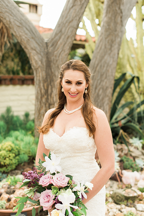 Orange county outdoor wedding at sherman library and gardens bride form fitting strapless lace gown with sweetheart neckline and pearl necklace holding white and pink floral bridal bouquet