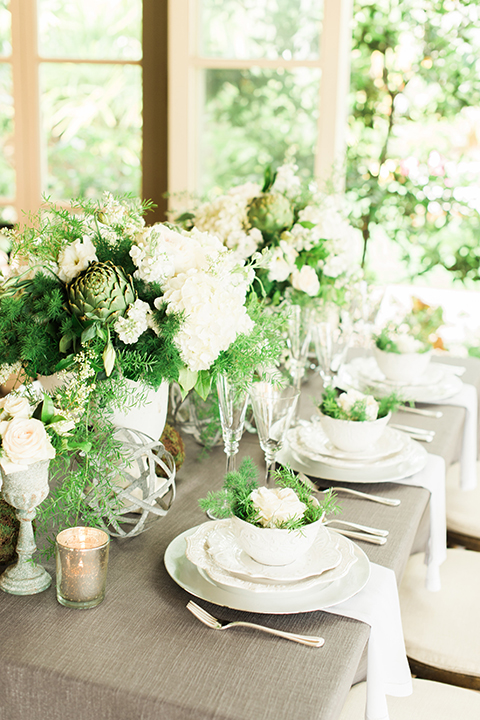 Orange county outdoor wedding shoot at sherman library and gardens table set up with light grey table linen and white place settings with green and white flower centerpiece decor with gold silverware and light brown and white wood chairs