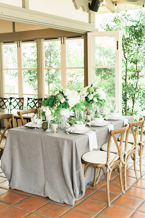 Orange county outdoor wedding shoot at sherman library and gardens table set up with light grey table linen and white place settings with green and white flower centerpiece decor with gold silverware and light brown and white wood chairs