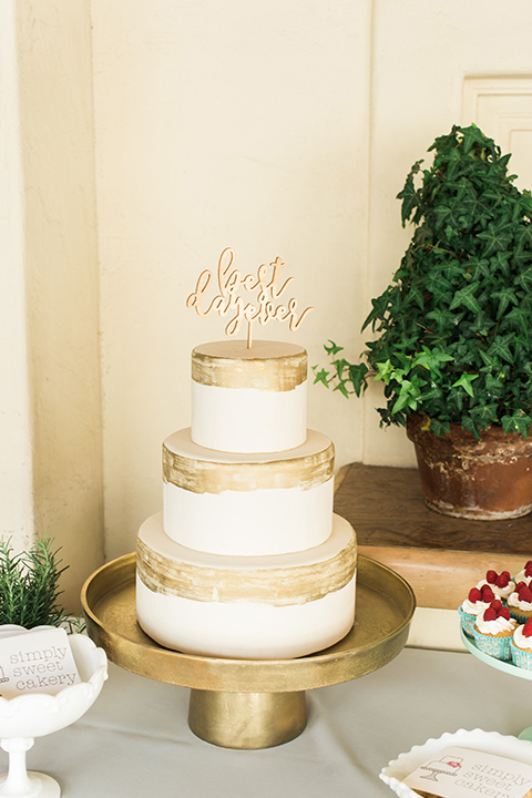 Orange county outdoor wedding shoot at sherman library and gardens table set up with light grey table linen and white place settings with green and white flower centerpiece decor with gold silverware and light brown and white wood chairs wedding cake three tier white and gold wedding cake with gold calligraphy cake topper