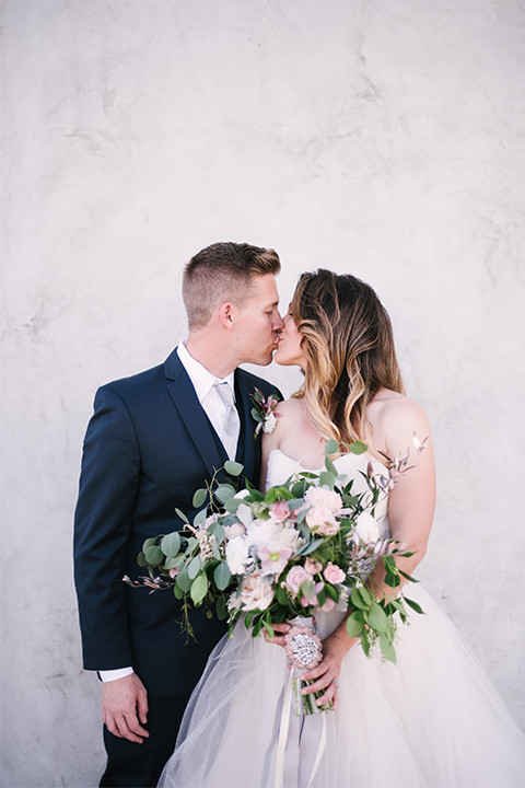 Orange county outdoor wedding at old ranch country club bride strapless ballgown with a sweetheart neckline and beaded detail with groom navy notch lapel suit with matching vest and white dress shirt with long silver tie and white floral boutonniere kissing and bride holding light pink and green floral bridal bouquet